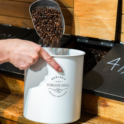 Person pouring coffee beans into a white container labeled 'Broèlerie Virgin Hill' on a wooden surface.