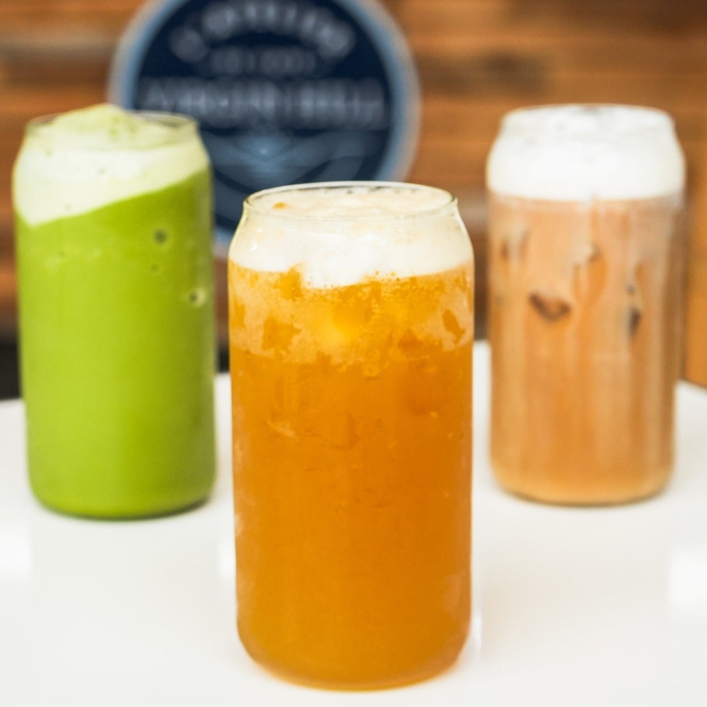 Three glasses of beer with foam, one green, one orange, and one brown, on a white surface with a blurred background.