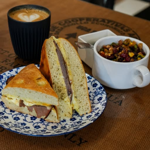 Half-eaten sandwich on a plate with a cup of coffee and a bowl of fruit on a wooden table.