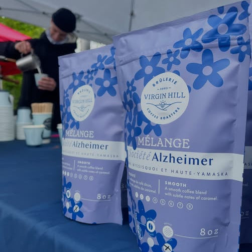 Two packages of Virgin Hill Mélange Café Alzheimer coffee on a blue tablecloth with a blurred background of people and tents.