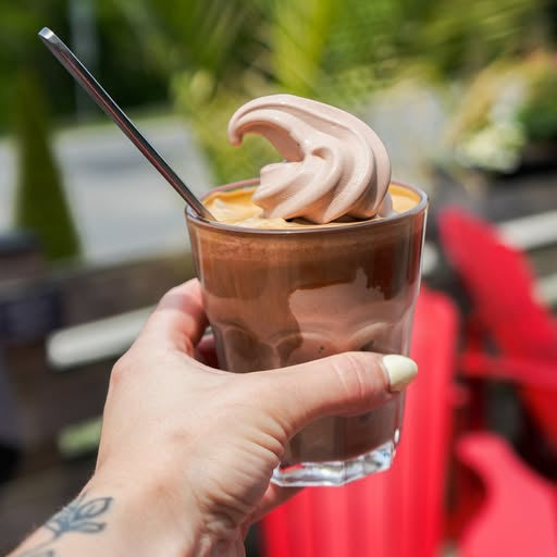 Hand holding a glass of iced coffee with a scoop of whipped cream outdoors.