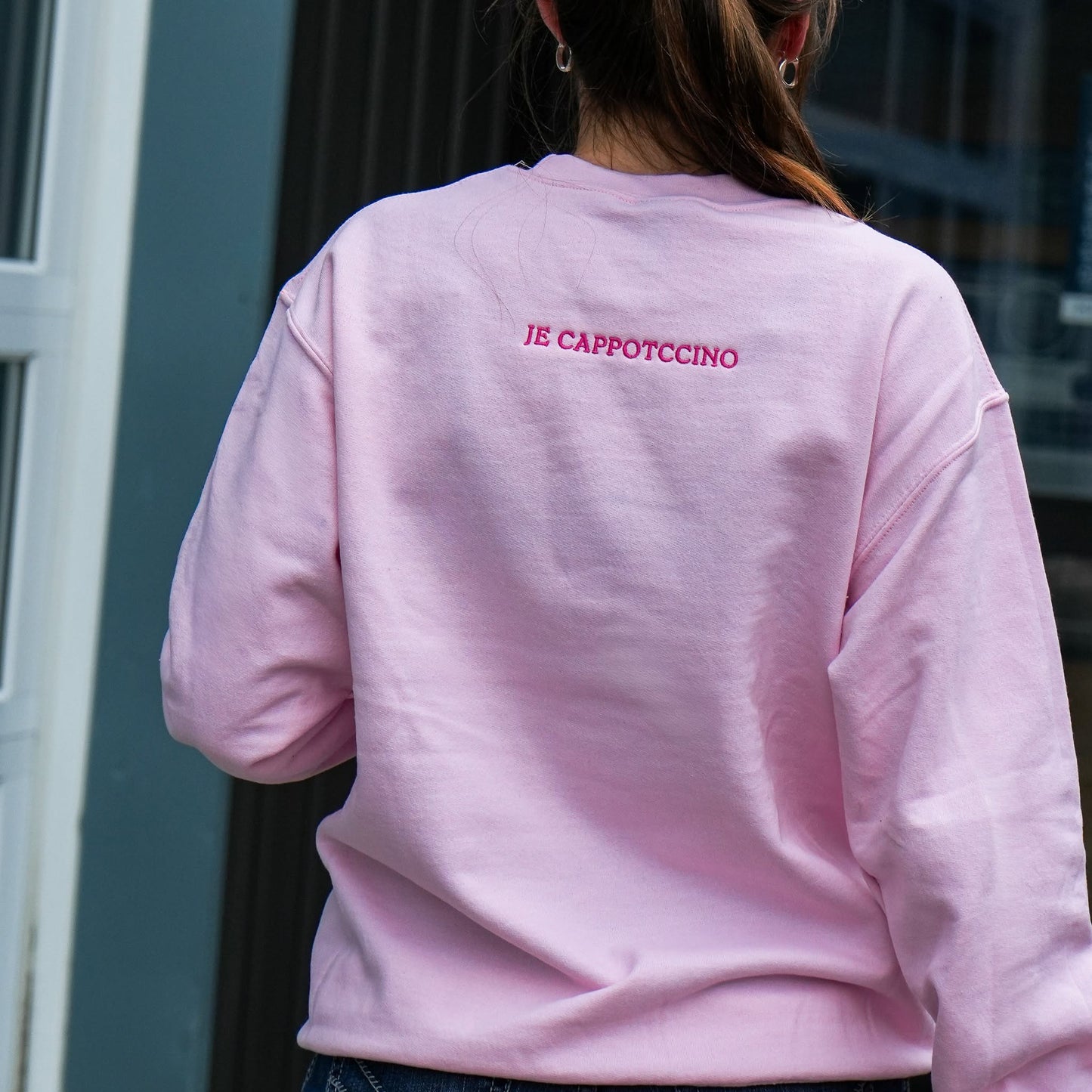 A person viewed from behind wearing a light pink crewneck sweatshirt with 'JE CAPPOTCCINO' embroidered in pink on the upper back, set against a dark gray building background.