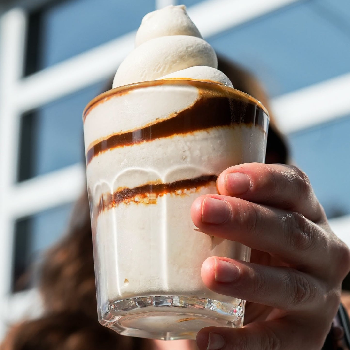 A close-up shot of a hand holding a clear glass cup filled with a layered creamy beverage topped with whipped cream and drizzles of dark chocolate sauce, with a blurred person and window background.