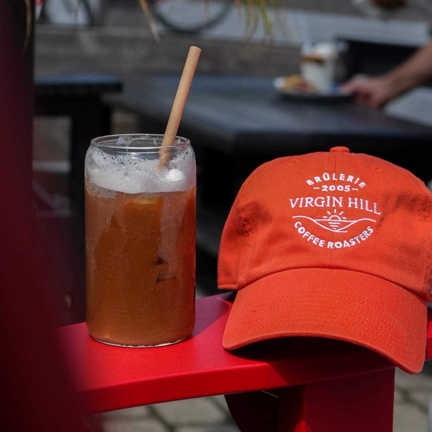 An orange-red baseball cap with white embroidered text reading 'Sölberie 2005 Virgin Hill Coffee Roasters' sits on a bright red table next to a glass of iced coffee with a straw in an outdoor cafe setting.