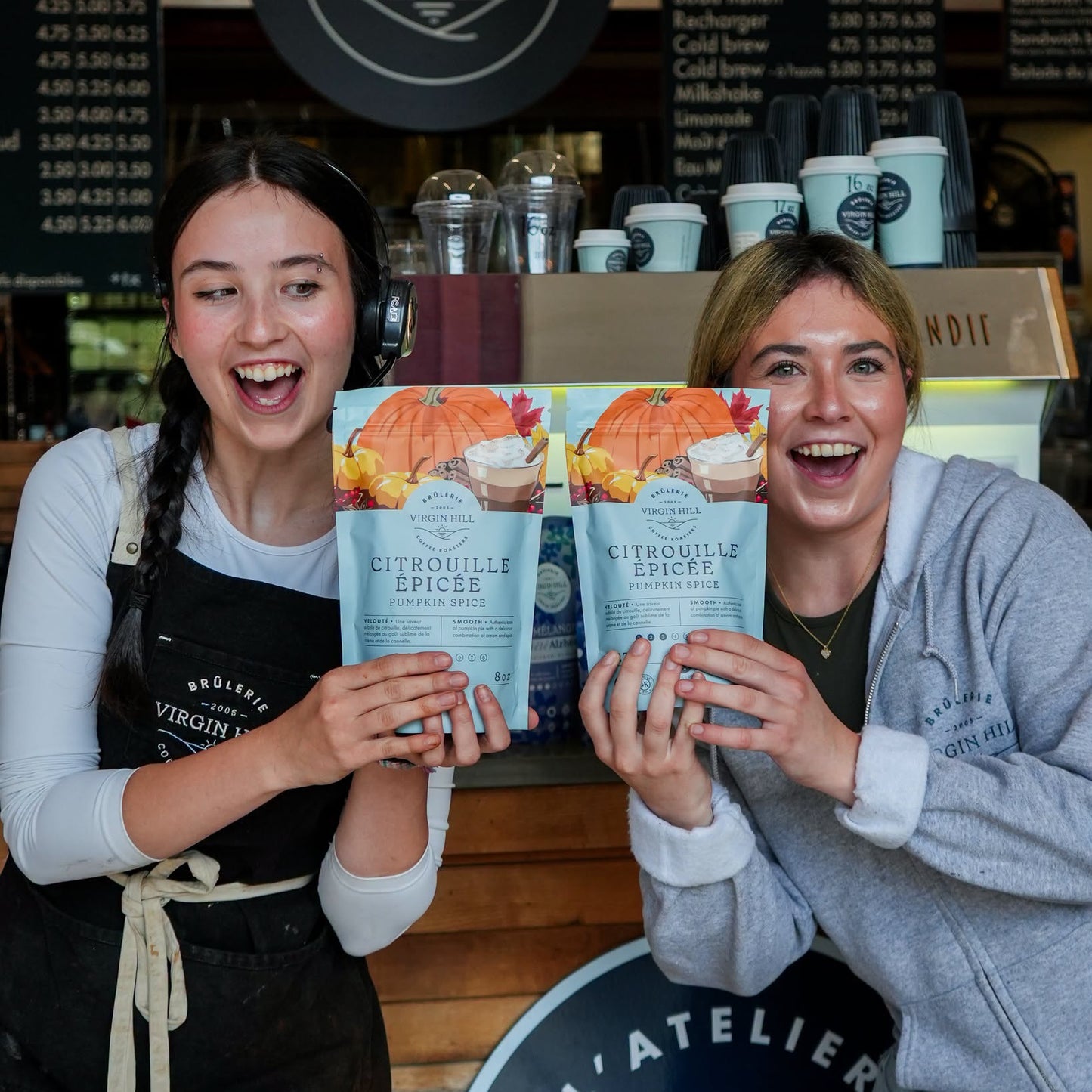 Two smiling women standing behind a cafe counter holding two boxes of Citrouille Spice Powder. The background shows a coffee menu board and cafe equipment.