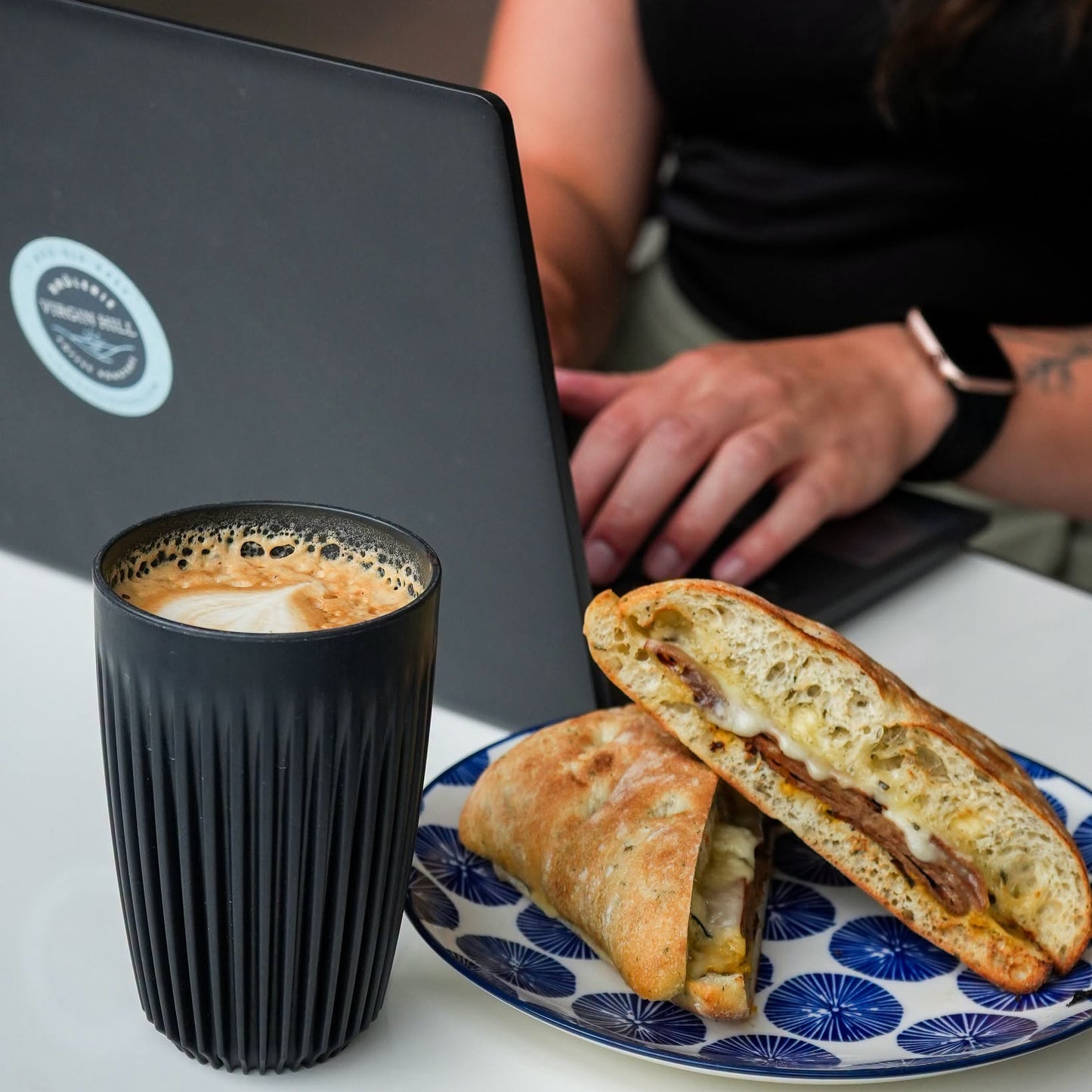 A lifestyle scene showing a black coffee cup with latte art, a plate with a toasted sandwich cut in half, and a black laptop with a circular sticker, with a person's hands and torso working on the laptop in the background.
