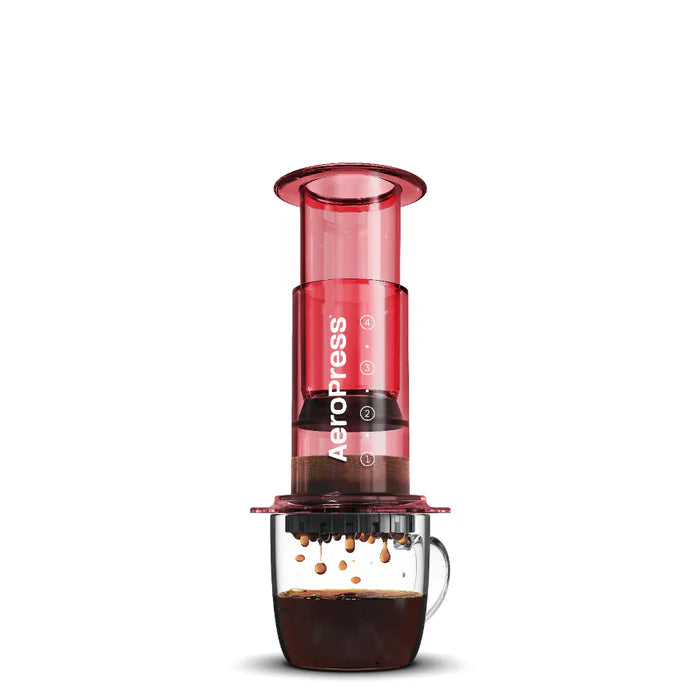 A red Aeropress clear coffee maker with a glass lower part and a plastic upper part, displayed against a white background.