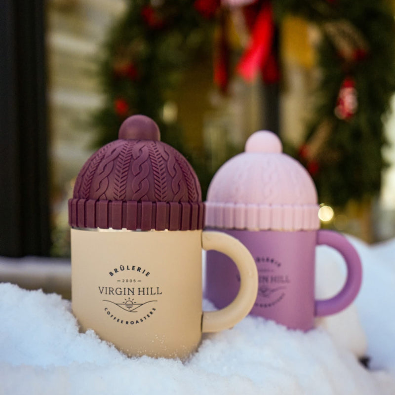 Two ceramic mugs with knit-style covers in the snow, one beige and one pink, with a blurred Christmas wreath in the background.