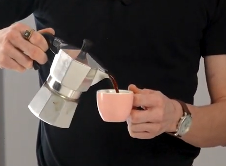 Person pouring coffee from a stovetop espresso maker into a pink cup.