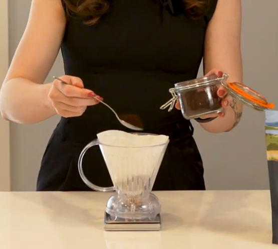 Person preparing coffee using a clear glass coffee maker and a small container.