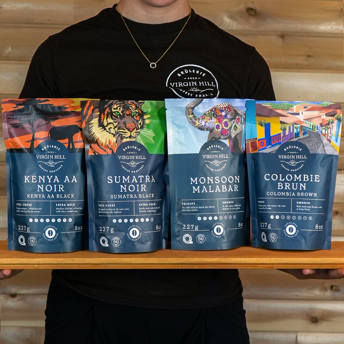 Person holding a wooden tray with coffee packages labeled 'Kenya AA Noir', 'Sumatra Noir', 'Monsoon Malabar', and 'Colombie Brun' from Virgin Hill Coffee.